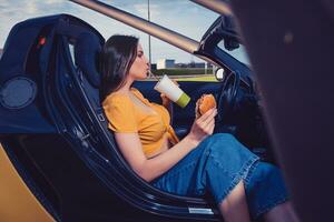 Lady in blue jeans and orange top is drinking beverage from paper cup, holding hamburger while sitting in yellow car cabrio. Fast food. Mock up photo