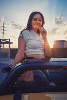 Young female in sunglasses and white t-shirt is eating pizza while posing standing in yellow car cabriolet. Fast food. Summer sunset. Close up photo