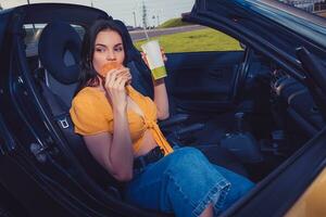 Model in blue jeans and orange top is eating hamburger and holding beverage in paper cup while sitting in yellow car cabriolet. Fast food. Copy space photo