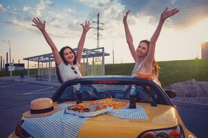 Ladies in casual outfit are smiling, raised up hands, posing in yellow car with french fries, pizza and soda on its trunk. Fast food. Copy space photo