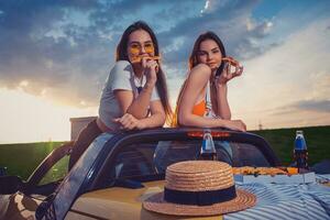 Charming girls eating pizza, posing in yellow car roadster with french fries, hat and soda in glass bottles on trunk. Fast food. Mock up photo