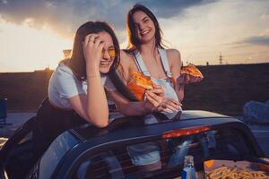 Two ladies in casual clothes eating pizza, laughing, posing in yellow car with french fries and soda in glass bottle on trunk. Fast food. Close up photo