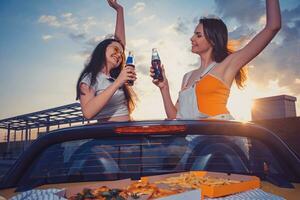 Two young girls smiling, cheering with soda in glass bottles, posing in yellow car cabrio with french fries and pizza on trunk. Fast food. Copy space photo
