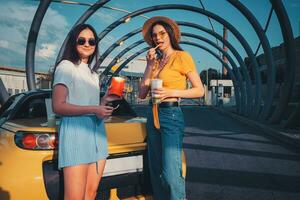 Young girls in casual clothes are smiling, enjoying french fries from paper package and beverages in paper cups, posing near yellow car. Copy space photo