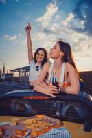 Happy females having fun and drinking soda in glass bottles, posing in yellow car with french fries and pizza on its trunk. Fast food. Copy space photo