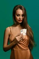 Brunette lady in silk beige dress. She looking at two playing cards in her hand, posing against green background. Poker, casino. Close-up photo