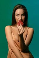 Young woman in silk golden dress. She is showing two red chips, posing against green background. Poker, casino. Close-up photo