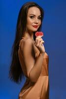 Young brunette female in silk beige dress. She smiling, showing two red chips, posing sideways on blue background. Poker, casino. Close-up photo