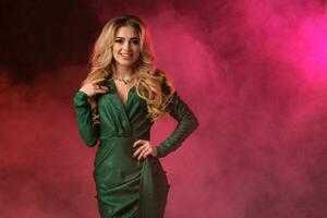 Blonde curly female in green stylish dress and jewelry. She put her hand on waist, smiling, posing against colorful smoky studio background. Close up photo