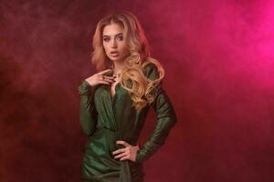 Alluring curly female in green stylish dress and jewelry. She has put her hand on waist, posing against colorful smoky studio background. Close up photo