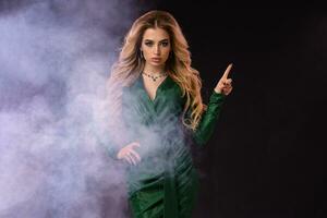 Blonde girl in green stylish dress and jewelry. Pointing at something by forefinger, posing on black smoky background. Close up, copy space photo