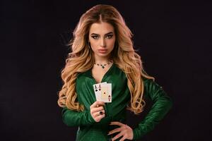 Blonde model in green stylish dress and jewelry. Put her hand on hip, showing two playing cards, posing on black background. Poker, casino. Close-up photo
