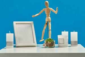 White table with green succulent in glass pot, wooden figurine of human, four candles, empty photo frame on it. Blue background. Close up