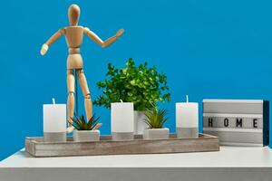 White table, green flower, succulent in pots, wooden figurine of human, candles in wooden stand, lamp with inscription home. Blue background. Close up photo