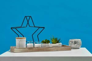 White table, wooden stand with green succulents in pots, two candles, jar. Black candlestick like iron star and silver one. Blue background. Close up photo