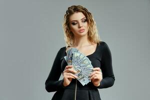 Blonde girl in black dress is holding some cash, posing against gray background. Gambling entertainment, poker, casino. Close-up. photo