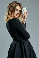 Blonde female in black dress showing two playing cards, posing sideways against gray background. Gambling entertainment, poker, casino. Close-up. photo