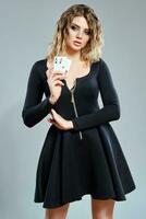 Blonde female in black short dress showing two playing cards, posing against gray background. Gambling entertainment, poker, casino. Close-up. photo