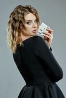 Blonde woman in black dress showing two playing cards, posing sideways against gray background. Gambling entertainment, poker, casino. Close-up. photo