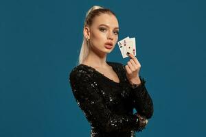 Blonde girl in black sequin dress showing two playing cards, posing against blue background. Gambling entertainment, poker, casino. Close-up photo