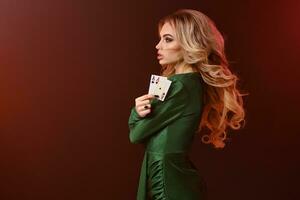 Blonde female in green dress and jewelry. Folded her hands, showing two playing cards, posing sideways on red background. Poker, casino. Close-up photo