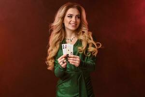 Blonde lady in green stylish dress and jewelry. Smiling and showing two aces, posing on colorful smoky background. Poker, casino. Close-up photo