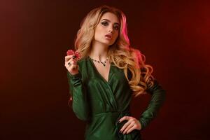 Blonde lady in green stylish dress and jewelry. She is showing two red chips, posing on red background with backlight. Poker, casino. Close-up photo