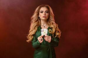 Curly blonde girl in green stylish dress and jewelry. Showing two playing cards, posing on colorful smoky background. Poker, casino. Close-up photo