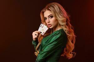 Blonde woman in green stylish dress and jewelry. Looking at you, showing two aces, posing sideways on brown background. Poker, casino. Close-up photo