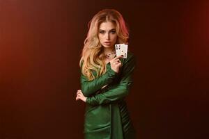 Blonde girl in green stylish dress and jewelry. Hands folded, showing two aces, posing against brown studio background. Poker, casino. Close-up photo