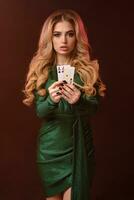 Blonde curly girl in green stylish dress and jewelry. She is showing two playing cards, posing on brown background. Poker, casino. Close-up photo