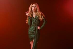 Blonde female with bright make-up, in green stylish dress and jewelry. Showing two red chips, posing on colorful background with backlight. Close-up photo