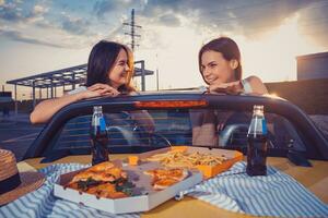 Young females are having fun, posing in yellow car roadster with french fries, pizza and soda in glass bottles on its trunk. Fast food. Mock up photo