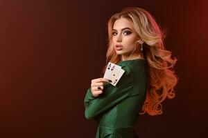 Blonde woman in green stylish dress and jewelry. Folded her hands, showing two aces, posing sideways on red studio background. Poker, casino. Close-up photo