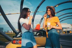Young females in casual outfit are smiling, eating french fries and holding beverage in paper cup while posing near yellow car. Fast food photo