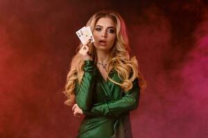 Blonde woman in green dress and jewelry. Showing two playing cards, opened her mouth, posing on colorful smoky background. Poker, casino. Close-up photo