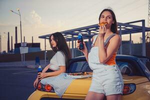 Attractive ladies in casual clothes eating pizza, enjoying soda water in glass bottles, posing near yellow car on parking lot. Close up, copy space photo