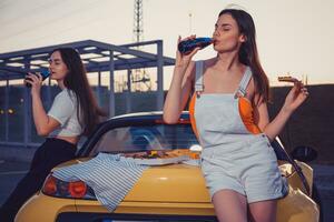 Females drinking carbonated beverage in glass bottles, eating pizza while posing near yellow car on parking lot. Fast food. Close up, mock up photo
