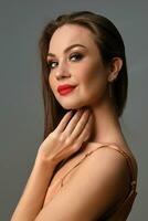 Brunette woman in silk beige dress. Smiling and touching her neck, posing standing sideways against gray background. Close up photo