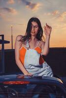 Sexy young female in white overall and orange t-shirt is posing outdoors standing in car cabriolet. Summer evening, sunset. Close up photo