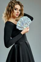 Maiden with curly hair, in black dress is holding some cash, posing sideways on gray background. Gambling entertainment, poker, casino. Close-up photo