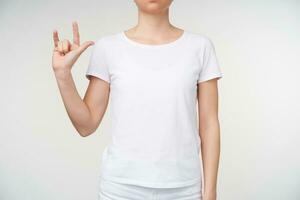 Studio photo of young woman keeping her hand raised and forming gesture while using sign language, standing over white background in casual wear