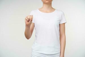 Studio photo of young female dressed in white t-shirt clenching her raised hand while forming letter m on sign language, isolated over white background
