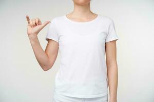 Horizontal photo of young woman dressed in casual wear keeping hand raised while showing letter y on sign language, isolated over white background