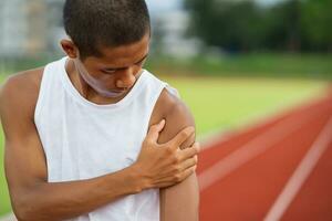 Athletes sport man runner wearing white sportswear to sitting feeling pain in his shoulder after practicing on a running track at a stadium, copy space. Runner sport injury concept. photo