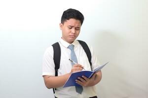 Indonesian senior high school student wearing white shirt uniform with gray tie writing on note book using pen with annoyed and frustrated expression. Isolated image on white background photo