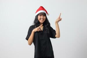 retrato de atractivo asiático mujer con rojo Papa Noel sombrero sensación feliz, señalando y demostración producto a el lado con dedo. nuevo año y Navidad concepto. aislado imagen en blanco antecedentes foto