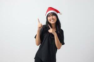 retrato de atractivo asiático mujer con rojo Papa Noel sombrero sensación feliz, señalando y demostración producto a el lado con dedo. nuevo año y Navidad concepto. aislado imagen en blanco antecedentes foto