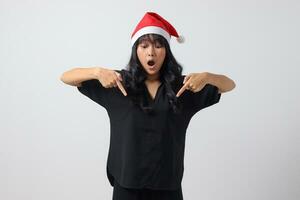 Portrait of attractive Asian woman with red Santa hat feeling happy, showing and pointing down with finger. New year and christmas concept. Isolated image on white background photo
