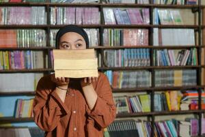 Portrait of Asian hijab woman covering her head with a pile of books in front of library bookshelf. Muslim girl reading a book. Concept of literacy and knowledge photo